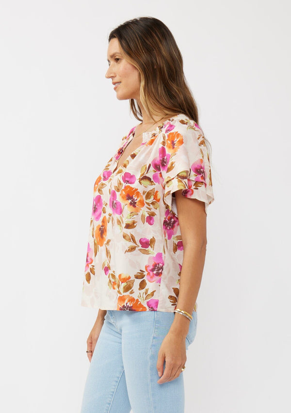 [Color: Natural/Orchid] A side facing image of a brunette model wearing a relaxed fit top in a floral print. With short double flutter sleeves, split v neckline, and a smocked detail at the neck. A dressy but casual floral top styled with light denim.  