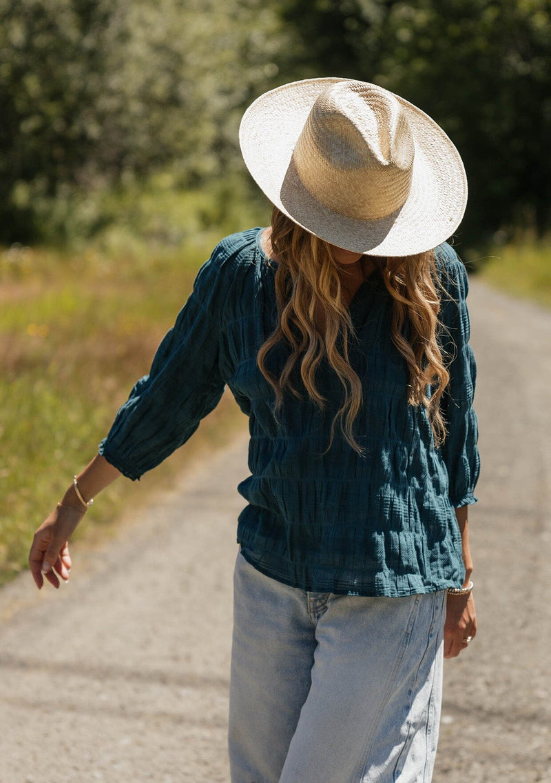 [Color: Deep Teal] A blonde model wearing a textured jacquard stripe blouse in teal. A relaxed fit blouse with a split v neckline and long sleeves. Styled with loose fit denim jeans.