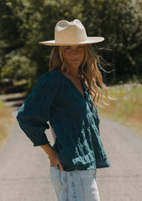 [Color: Deep Teal] A blonde model wearing a textured jacquard stripe blouse in teal. A relaxed fit blouse with a split v neckline and long sleeves. Styled with loose fit denim jeans. 