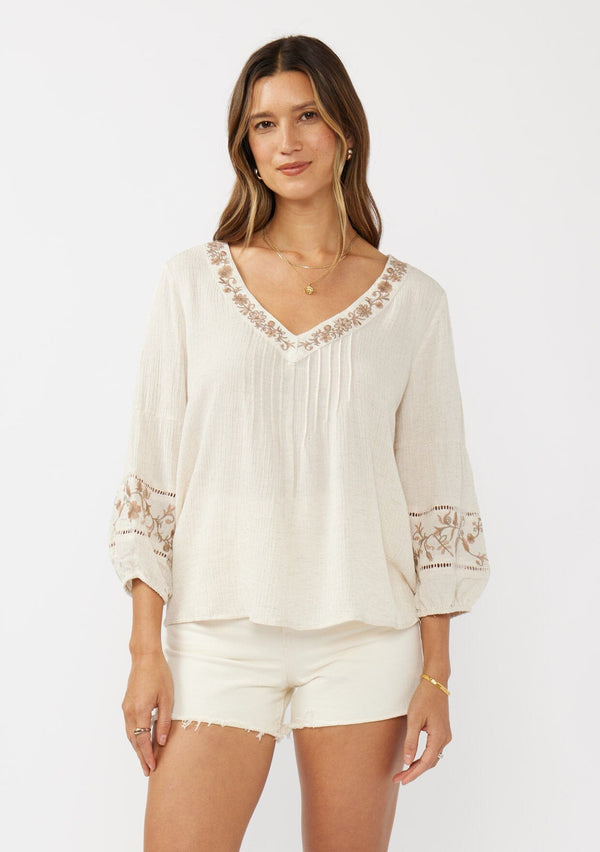 [Color: Natural/Brown] An image of a brunette model wearing an ultra bohemian blouse with embroidered details. A beige natural tone blouse with a v neckline, floral embroidery, and long sleeves with elastic cuffs. A relaxed fit boho top paired with neutral off white shorts.