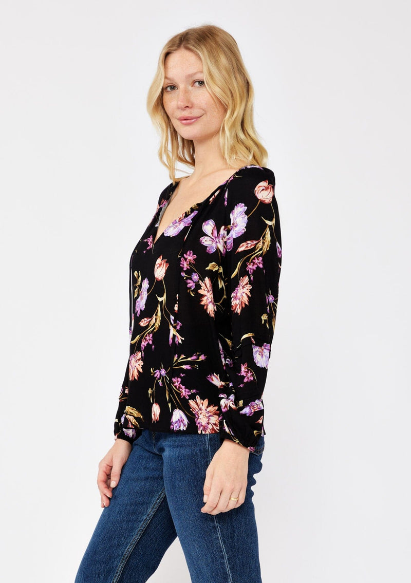 [Color: Black/Purple] A blonde model wearing a flowy bohemian top in a black and purple floral print. Featuring long sleeves with pintuck details and a split v neckline with ties. A casual, everyday top, perfect for any occasion during the Fall season. 