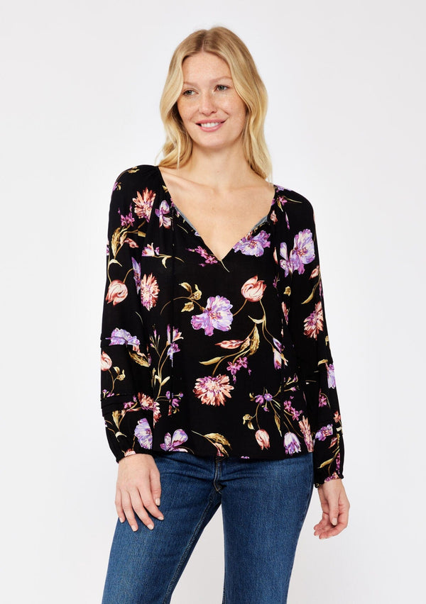 [Color: Black/Purple] A blonde model wearing a flowy bohemian top in a black and purple floral print. Featuring long sleeves with pintuck details and a split v neckline with ties. A casual, everyday top, perfect for any occasion during the Fall season. 