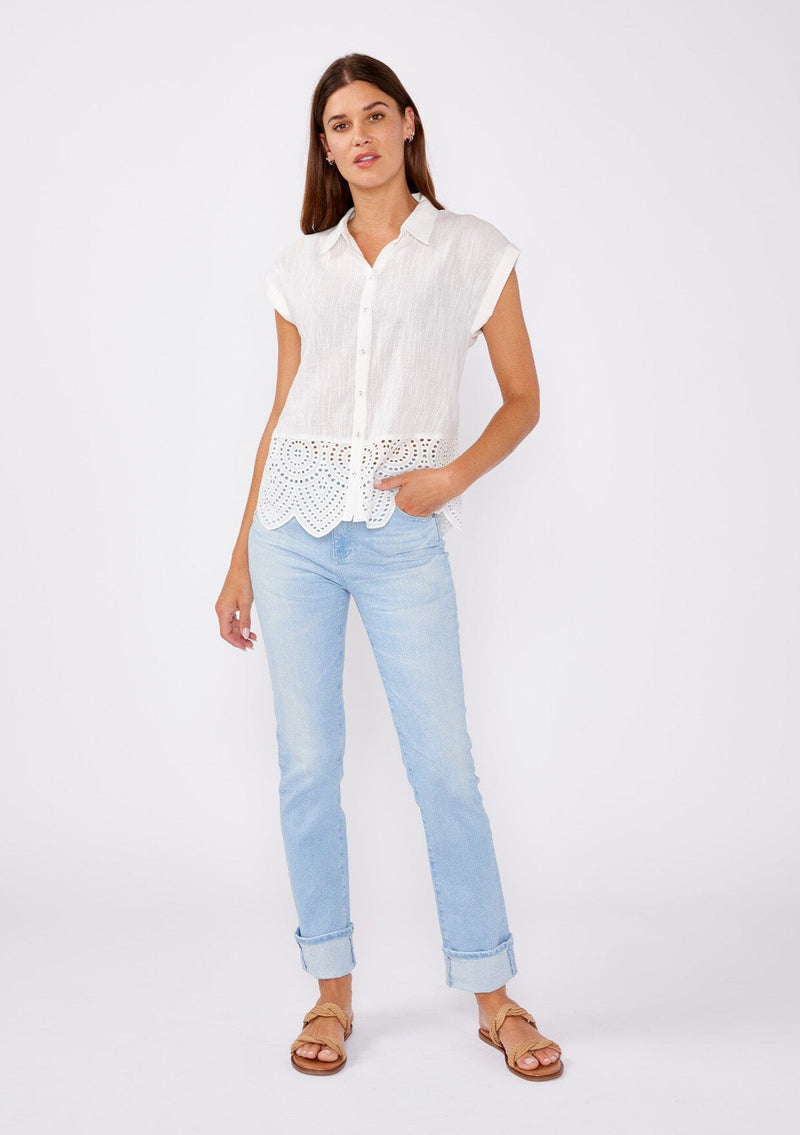 [Color: White] A brunette model wearing a breezy cotton camp shirt with a unique embroidered eyelet design and scalloped edge. This white collared top features a functional button front, cap sleeves, and a relaxed fit. Paired with light wash denim jeans for that effortless summer style.