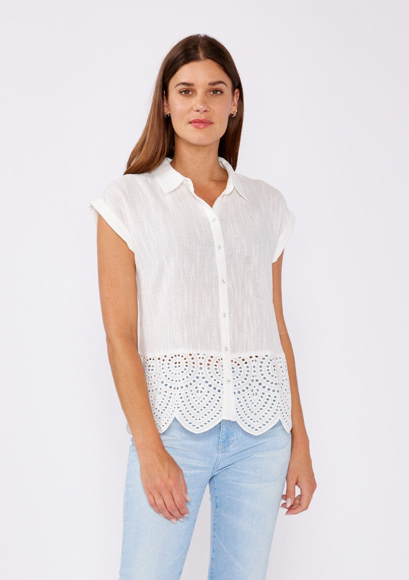 [Color: White] A brunette model wearing a breezy cotton camp shirt with a unique embroidered eyelet design and scalloped edge. This white collared top features a functional button front, cap sleeves, and a relaxed fit. Paired with light wash denim jeans for that effortless summer style. 