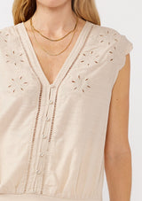 [Color: Buff] A detail image of a blonde model wearing a taupe rayon and cotton blend top with an embroidered floral cutout design. This casual spring top features a v neckline, button front, short dolman sleeves, and a hip hugging elastic hem at the back. 