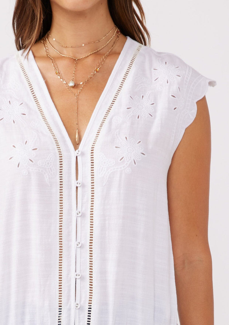 [Color: White] A detail shot of a brunette model wearing a rayon and cotton blend top with an embroidered floral cutout design. This casual white top features a v neckline, button front, short dolman sleeves, and a hip hugging elastic hem at the back. 