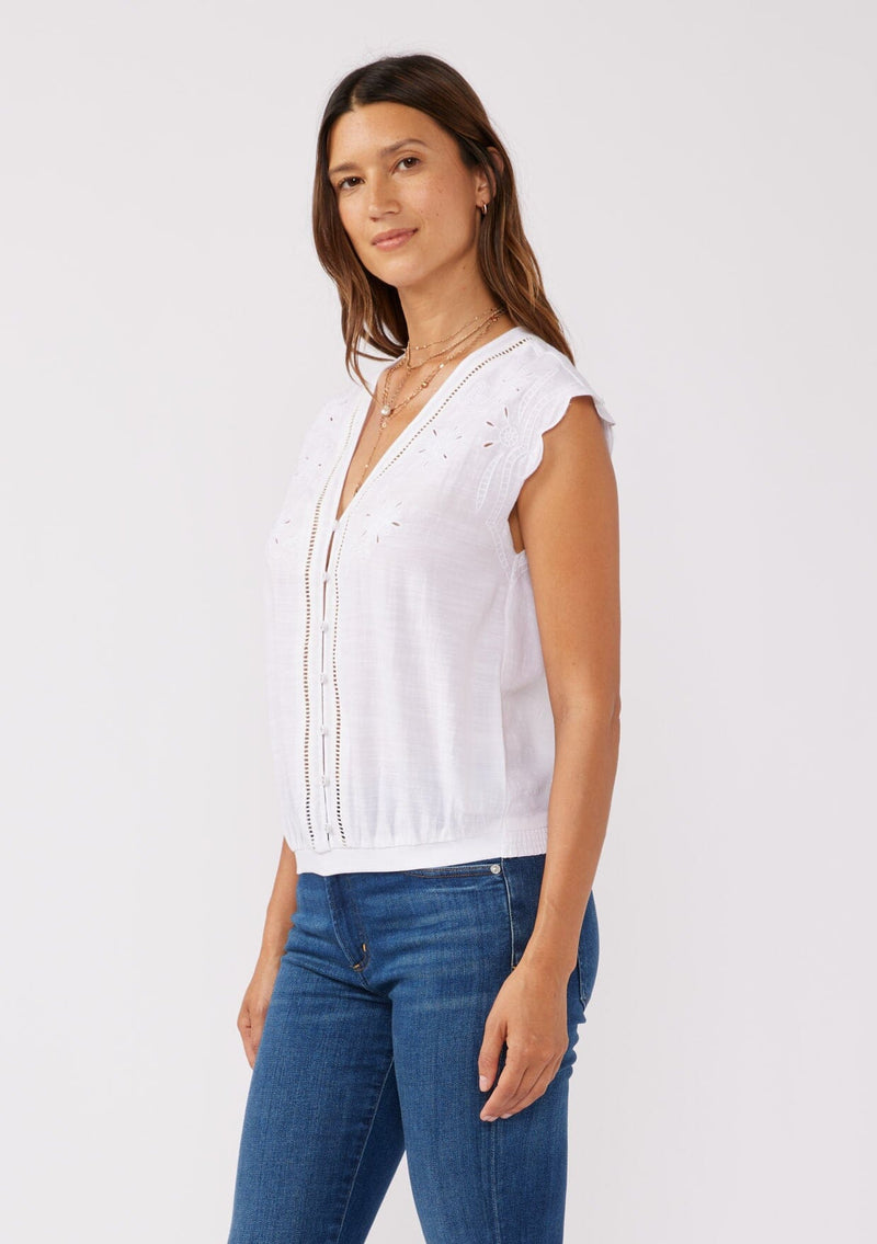 [Color: White] A side facing image of a brunette model wearing a rayon and cotton blend top with an embroidered floral cutout design. This casual white top features a v neckline, button front, short dolman sleeves, and a hip hugging elastic hem at the back. 