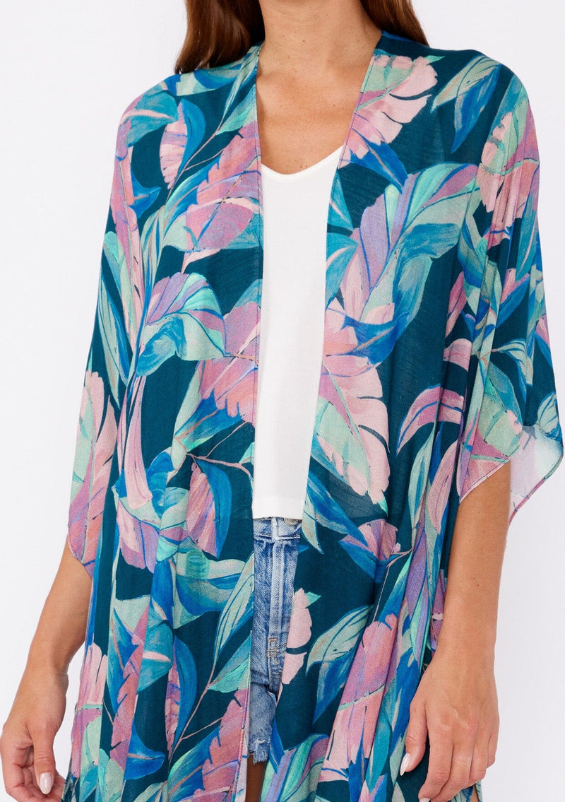 [Color: Teal/Dusty Orchid] A brunette model wearing a blue and pink tropical inspired palm leaf print kimono. A lightweight kimono perfect for the beach or to style a casual outfit. With an open front, mid length , half kimono sleeves, and side vents.