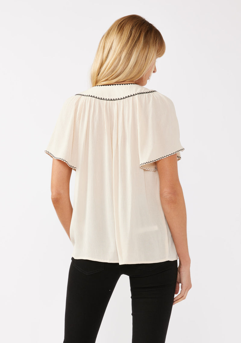 [Color: Shell/Black] A blonde model wearing a classic bohemian top in off white. With an embroidered blanket stitch trim, short flutter sleeves, and a split v neckline. Perfect for any wardrobe; styled with black denim for a cool and casual look.  