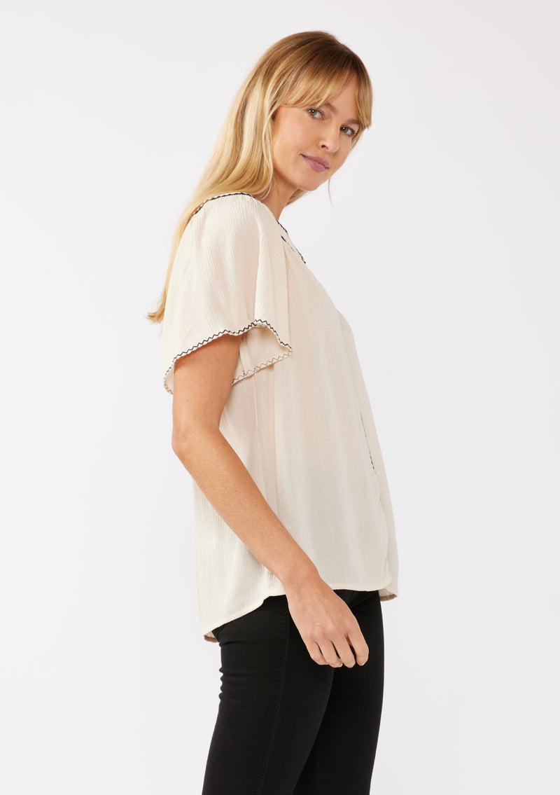 [Color: Shell/Black] A blonde model wearing a classic bohemian top in off white. With an embroidered blanket stitch trim, short flutter sleeves, and a split v neckline. Perfect for any wardrobe; styled with black denim for a cool and casual look.  