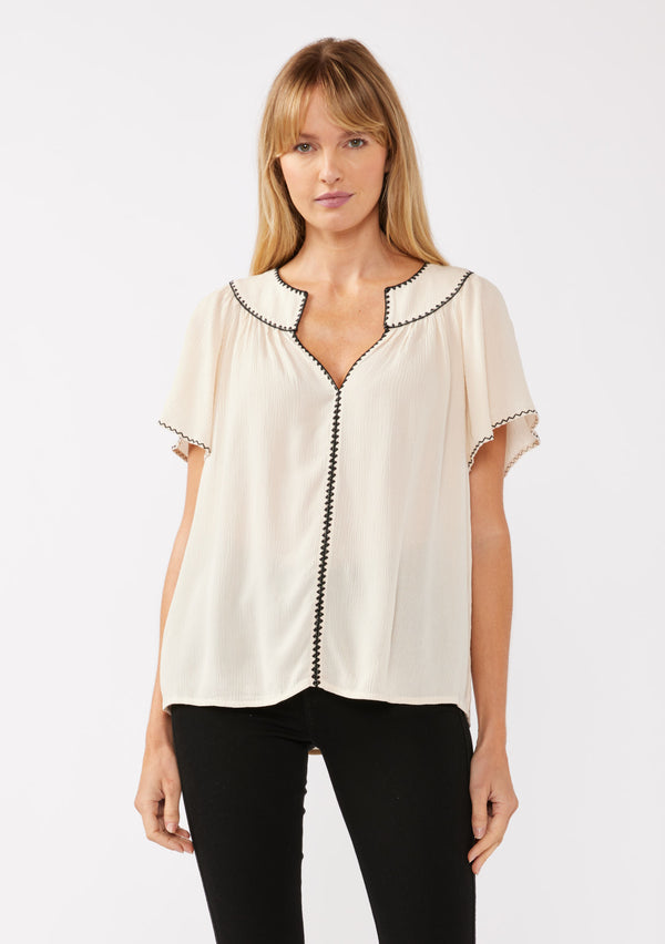 [Color: Shell/Black] A blonde model wearing a classic bohemian top in off white. With an embroidered blanket stitch trim, short flutter sleeves, and a split v neckline. Perfect for any wardrobe; styled with black denim for a cool and casual look.  