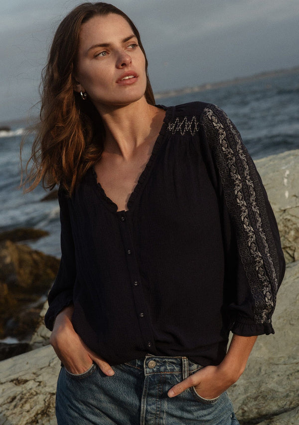 [Color: Navy] A front facing image of a brunette model wearing an embroidered, navy blue blouse with a button front, ruffle trimmed v-neckline, 3/4 sleeves, and elastic wrist cuffs. Paired with denim bottoms for a laid back look.