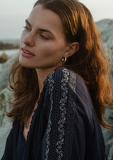 [Color: Navy] A detail image of a brunette model wearing an embroidered, navy blue blouse with a button front, ruffle trimmed v-neckline, 3/4 sleeves, and elastic wrist cuffs. Paired with denim bottoms for a laid back look. 