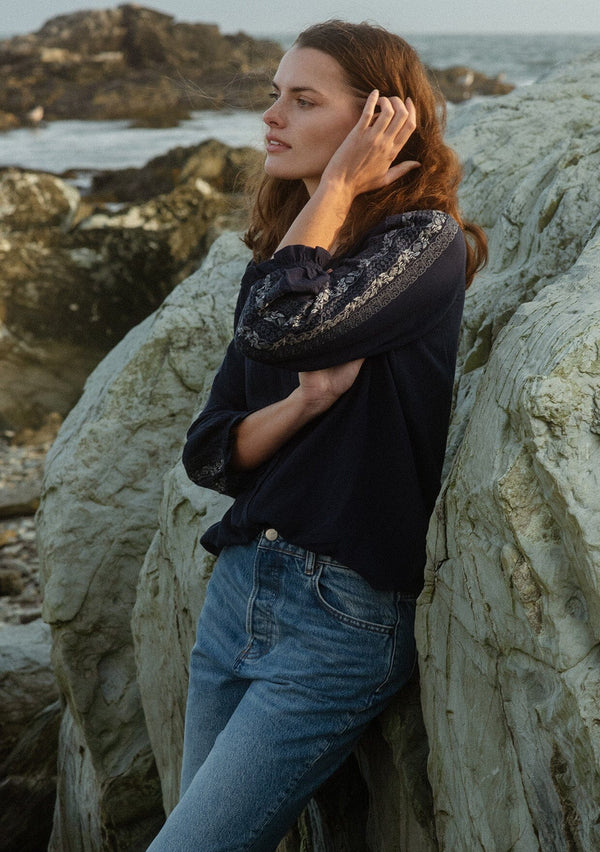 [Color: Navy] A side facing image of a brunette model wearing an embroidered, navy blue blouse with a button front, ruffle trimmed v-neckline, 3/4 sleeves, and elastic wrist cuffs. Paired with denim bottoms for a laid back look.