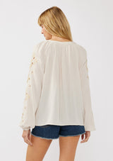 [Color: Natural] A blonde model wearing a viscose and linen blend peasant blouse with a contrast embroidery with cutout details. This split v-neckline blouse with long sleeves and elastic cuffs is the perfect casual piece for a relaxed resort look. 