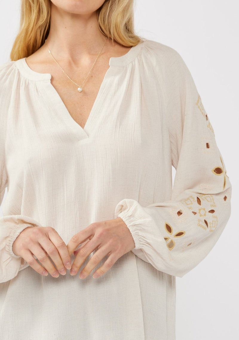 [Color: Natural] A detail image of a blonde model wearing a viscose and linen blend peasant blouse with a contrast embroidery with cutout details. This split v-neckline blouse with long sleeves and elastic cuffs is the perfect casual piece for a relaxed resort look. 