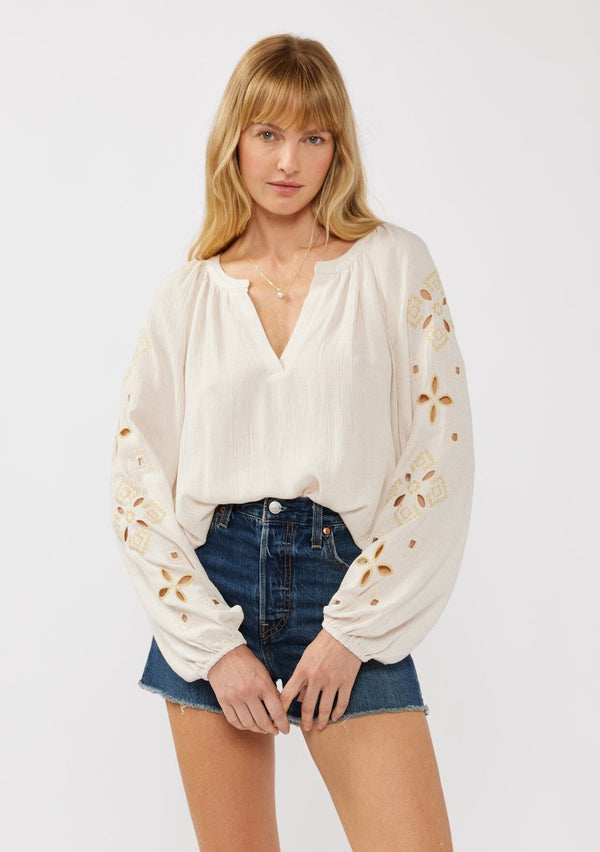 [Color: Natural] A blonde model wearing a viscose and linen blend peasant blouse with a contrast embroidery with cutout details. This split v-neckline blouse with long sleeves and elastic cuffs is the perfect casual piece for a relaxed resort look. 