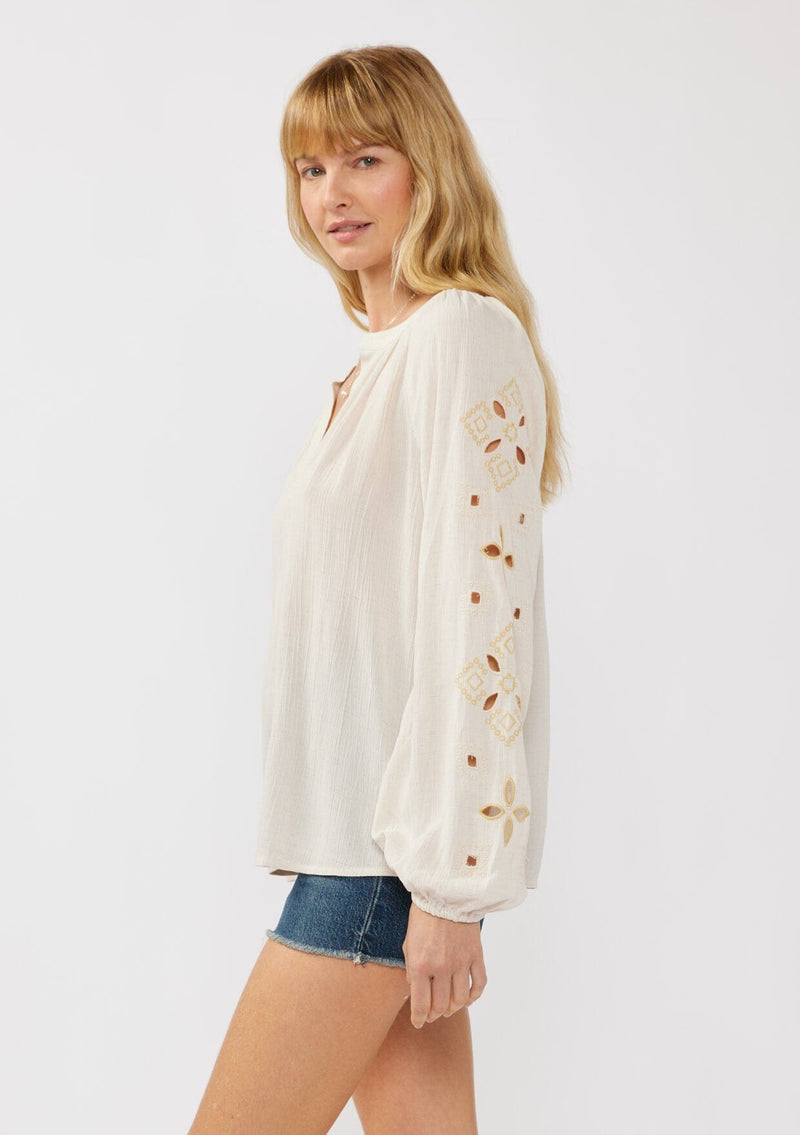 [Color: Natural] A blonde model wearing a viscose and linen blend peasant blouse with a contrast embroidery with cutout details. This split v-neckline blouse with long sleeves and elastic cuffs is the perfect casual piece for a relaxed resort look. 