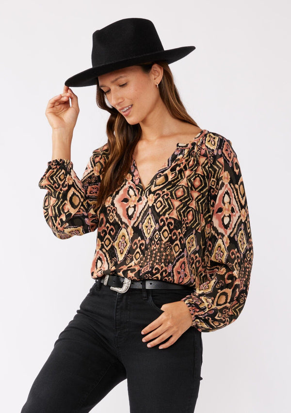 [Color: Grey/Dusty Rose] A brunette model wearing an ultra boho blouse with geometric ikat print in dusty pink and yellow. A casual fall blouse with a split v-neckline, long sleeves, and a relaxed fit. Styled with all black accessories for a laid-back look. 