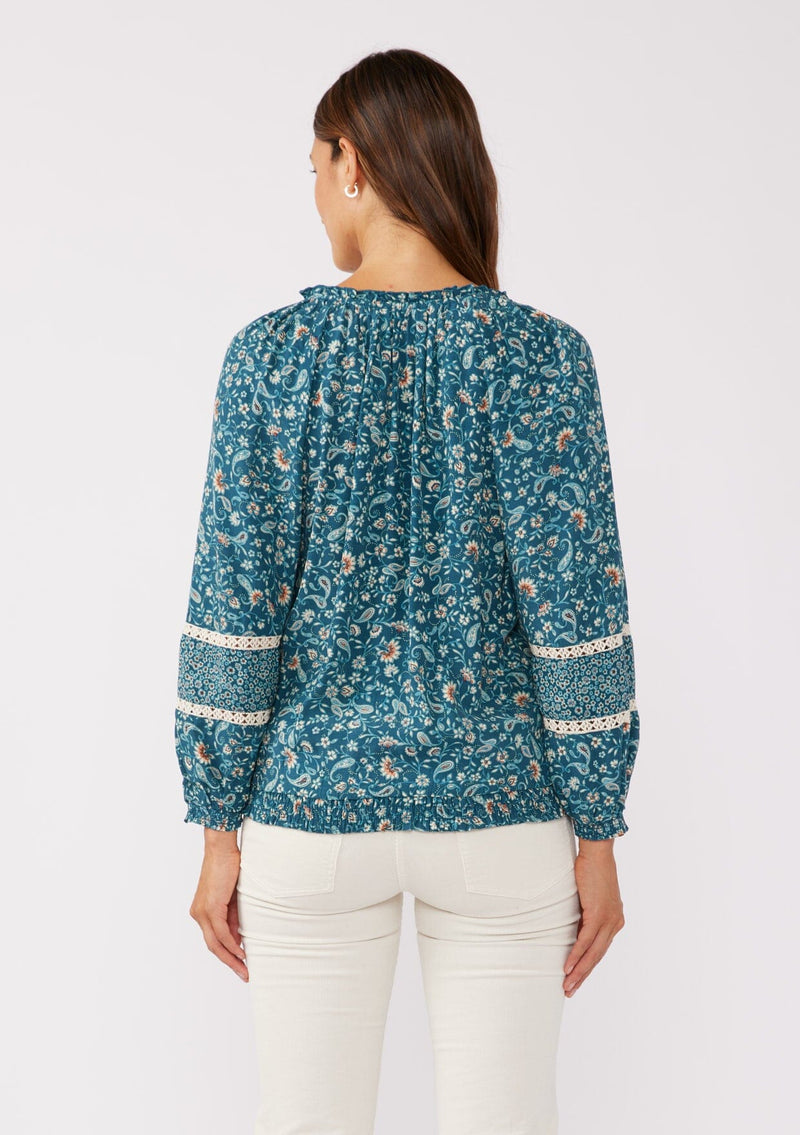 [Color: Teal/Cream] A back facing image of a brunette model wearing a paisley floral print blouse. With a split v-neckline with ties, long sleeves, lace trim inserts, and a smocked elastic hemline. A feminine boho top with a relaxed fit styled with of white corduroy bottoms. 