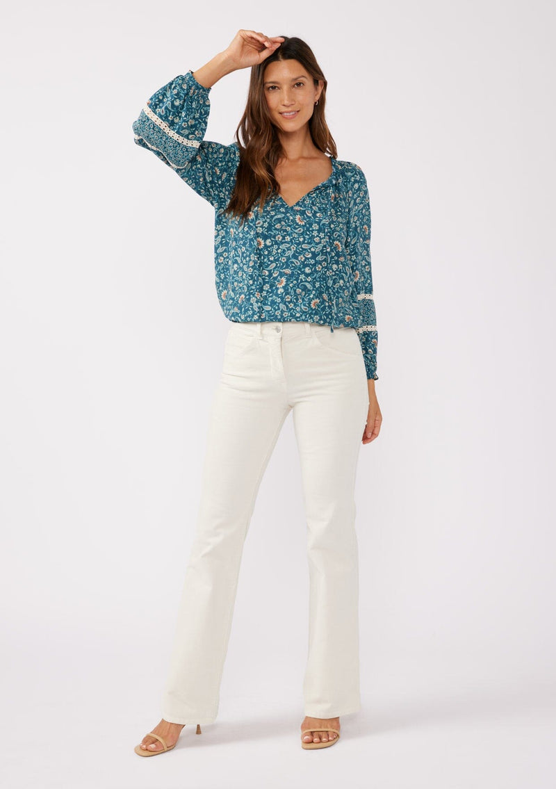 [Color: Teal/Cream] A full body front facing image of a brunette model wearing a paisley floral print blouse. With a split v-neckline with ties, long sleeves, lace trim inserts, and a smocked elastic hemline. A feminine boho top with a relaxed fit styled with of white corduroy bottoms. 