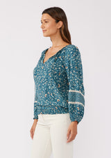 [Color: Teal/Cream] A side facing image of a brunette model wearing a paisley floral print blouse. With a split v-neckline with ties, long sleeves, lace trim inserts, and a smocked elastic hemline. A feminine boho top with a relaxed fit styled with of white corduroy bottoms. 