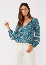 [Color: Teal/Cream] A front facing image of a brunette model wearing a paisley floral print blouse. With a split v-neckline with ties, long sleeves, lace trim inserts, and a smocked elastic hemline. A feminine boho top with a relaxed fit styled with of white corduroy bottoms. 