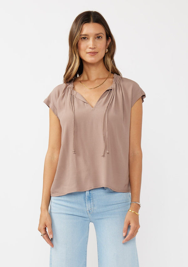 [Color: Mocha] A brunette model wearing a relaxed fit  brown mocha top. With self neck ties, ruched split v neckline, and short dolman sleeves. Styled with a cute wide leg denim for a laid back look. 