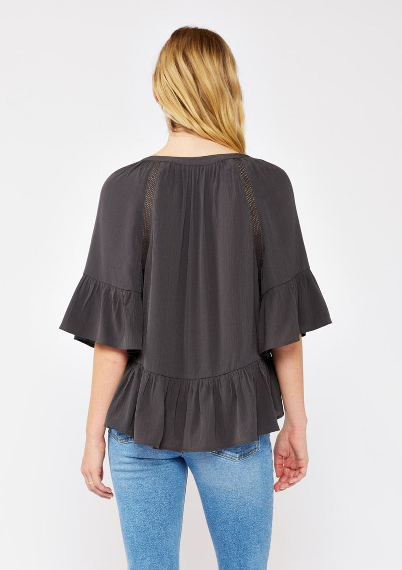 [Color: Charcoal] A blonde model wearing a grey relaxed fit peplum style blouse. A button front top with flutter half sleeves, split v neckline, and lace trim inserts. Styled with light denim jeans for an ultra boho look.