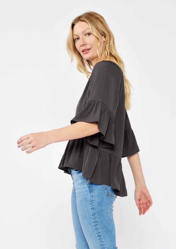 [Color: Charcoal] A blonde model wearing a grey relaxed fit peplum style blouse. A button front top with flutter half sleeves, split v neckline, and lace trim inserts. Styled with light denim jeans for an ultra boho look.