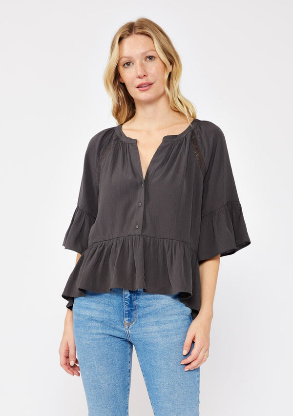 [Color: Charcoal] A blonde model wearing a grey relaxed fit peplum style blouse. A button front top with flutter half sleeves, split v neckline, and  lace trim inserts. Styled with light denim jeans for an ultra boho look. 