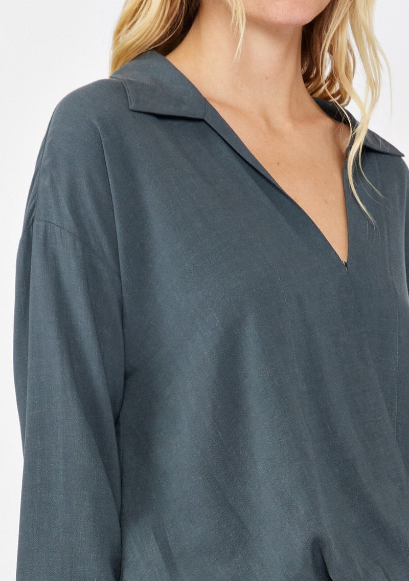 [Color: Steel Blue] A blonde model wearing a grey blue blouse in a soft linen blend. With a collared neckline, flattering v neckline, front hook and eye closure, and long sleeves with button cuffs. Completed with a comfortable half elastic waist at the back for a comfortable fit.