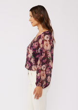 [Color: Plum/Dusty Rose] A side facing brunette model wearing a purple floral chiffon top with pleated long sleeves, button front, split v-neckline, and elastic hem. Perfect for holidays, work events, or date nights.