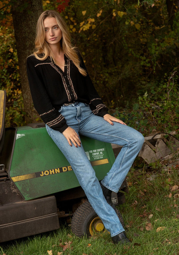 [Color: Black/Mocha] A front facing image of a blonde model wearing a black button-front blouse with contrasting embroidered details throughout. The boho blouse features a relaxed fit, long sleeves, a split V-neckline, and button-cuff closures. Styled with denim for the perfect fall ensemble. 