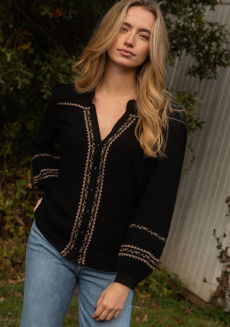 [Color: Black/Mocha] A front facing image of a blonde model wearing a black button-front blouse with contrasting embroidered details throughout. The boho blouse features a relaxed fit, long sleeves, a split V-neckline, and button-cuff closures. Styled with denim for the perfect fall ensemble. 