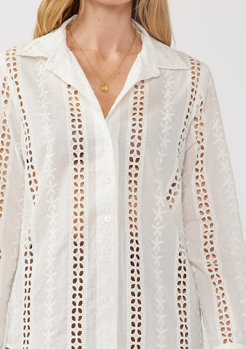 [Color: White] A detail image of a blonde model wearing floral eyelet shirt with a collared neckline, button front, and long sleeves with button cuffs. A sophisticated spring top paired with brown denim bottoms for spring. 