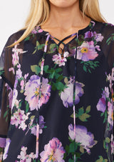 [Color: Navy/Lilac] A detail image of a blonde model wearing a navy and purple floral print blouse crafted in lightweight chiffon. This boho chic blouse features a split v-neckline with a lace up front, long sleeves, and a relaxed fit. Perfect for special occasions and dinner dates. 