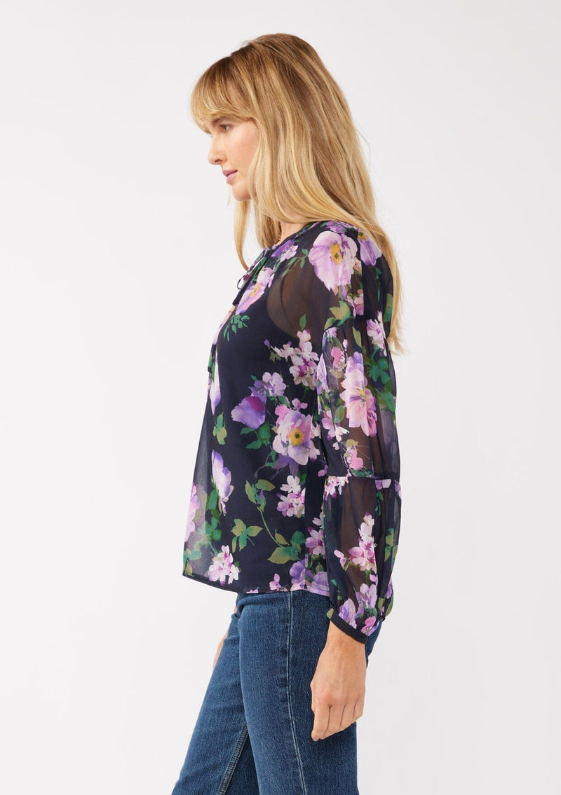 [Color: Navy/Lilac] A side facing image of a blonde model wearing a navy and purple floral print blouse crafted in lightweight chiffon. This boho chic blouse features a split v-neckline with a lace up front, long sleeves, and a relaxed fit. Perfect for special occasions and dinner dates. 