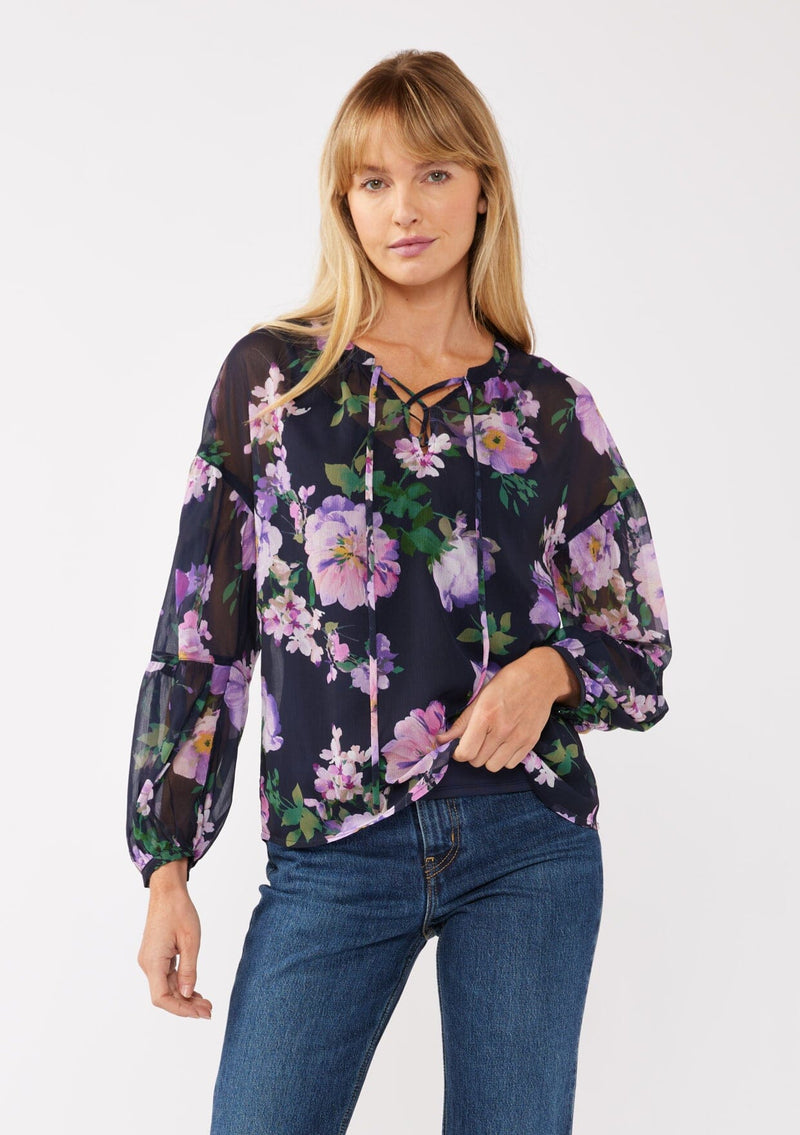 [Color: Navy/Lilac] A front facing image of a blonde model wearing a navy and purple floral print blouse crafted in lightweight chiffon. This boho chic blouse features a split v-neckline with a lace up front, long sleeves, and a relaxed fit. Perfect for special occasions and dinner dates. 