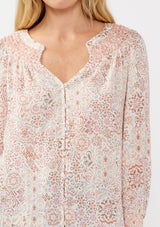 [Color: Natural/Dusty Pink] A detail image of a blond model wearing a classic v-neck bohemian blouse with a pink abstract print, smocked neckline and cuffs, and button front. 