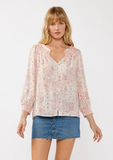 [Color: Natural/Dusty Pink] A front facing image of a blond model wearing a classic v-neck bohemian blouse with a pink abstract print, smocked neckline and cuffs, and button front. 