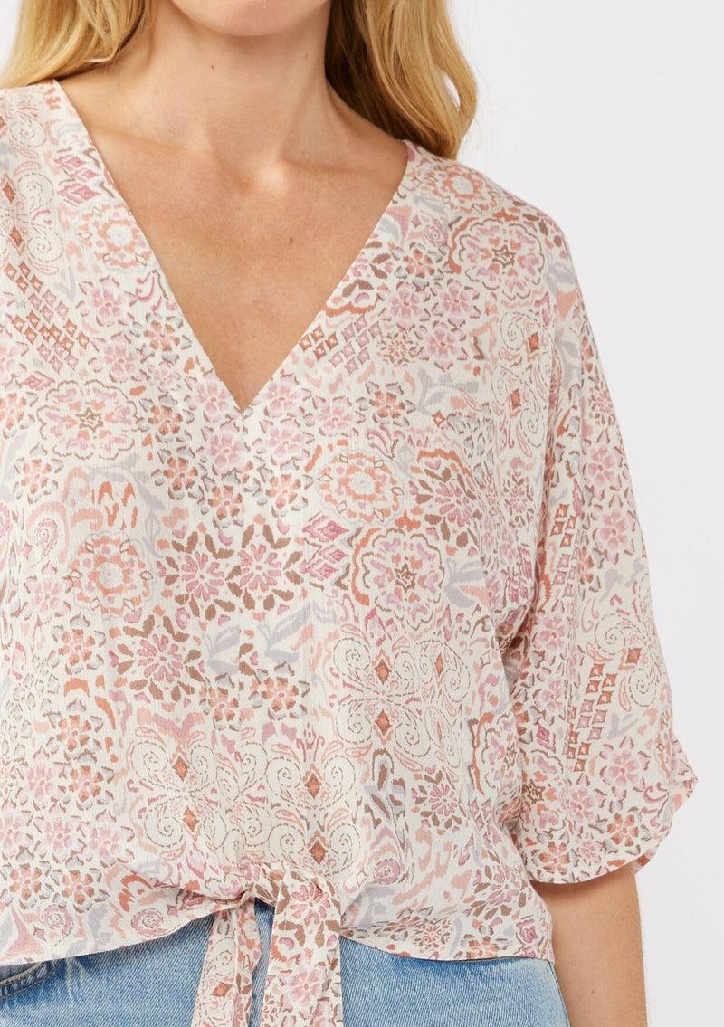 [Color: Natural/Dusty Pink] A detail image of a blonde model wearing a pink bohemian print blouse with a v-neckline, tie front detail at the hem, and flowy kimono sleeves. Perfect for resort styling and casual outings. 