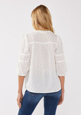 [Color: White] A back-facing image of a blonde model wearing a bohemian white blouse featuring a stripe clip jacquard pattern. This lightweight blouse includes a pintuck yoke, 3/4 length sleeves with tiered details, and a flattering split v-neckline. Ideal for casual outings, it’s a must-have addition to any boho wardrobe.