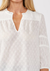 [Color: White] A detail image of a blonde model wearing a bohemian white blouse featuring a stripe clip jacquard pattern. This lightweight blouse includes a pintuck yoke, 3/4 length sleeves with tiered details, and a flattering split v-neckline. Ideal for casual outings, it’s a must-have addition to any boho wardrobe.