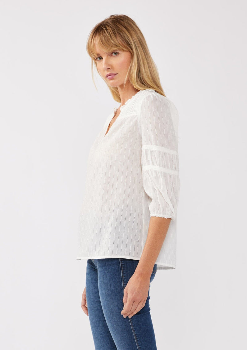 [Color: White] A side-facing image of a blonde model wearing a bohemian white blouse featuring a stripe clip jacquard pattern. This lightweight blouse includes a pintuck yoke, 3/4 length sleeves with tiered details, and a flattering split v-neckline. Ideal for casual outings, it’s a must-have addition to any boho wardrobe.