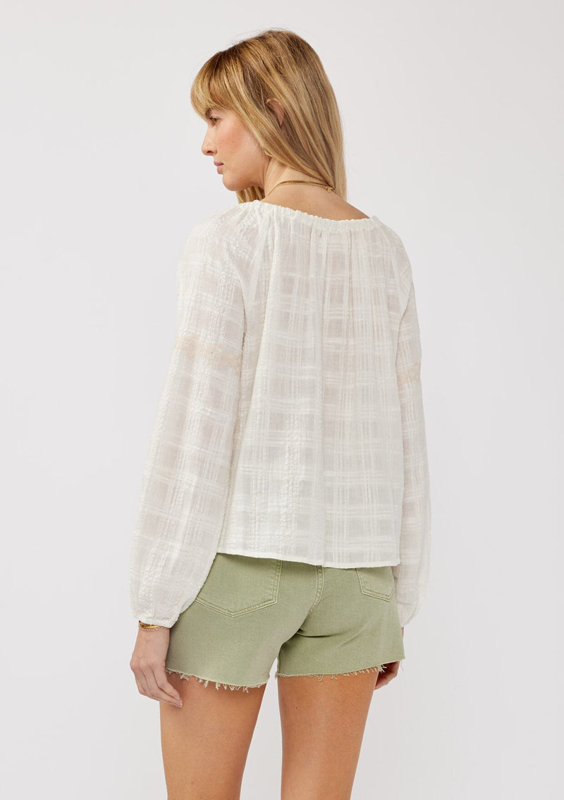 [Color: White/Taupe] A back facing image of a blonde model wearing a white boho blouse crafted in a textured plaid seersucker with an elastic neckline, tie neck detail, long sleeves, lace trim inserts, and contrast embroidery. 