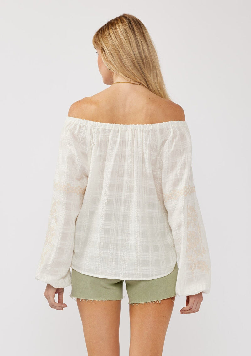 [Color: White/Taupe] A back facing image of a blonde model wearing an off shoulder top crafted in a textured plaid seersucker with an elastic neckline, tie neck detail, long sleeves, lace trim inserts, and contrast embroidery. 