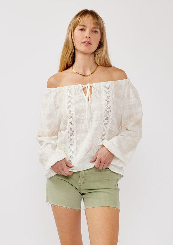 [Color: White/Taupe] A front facing image of a blonde model wearing an off shoulder top crafted in a textured plaid seersucker with an elastic neckline, tie neck detail, long sleeves, lace trim inserts, and contrast embroidery. 