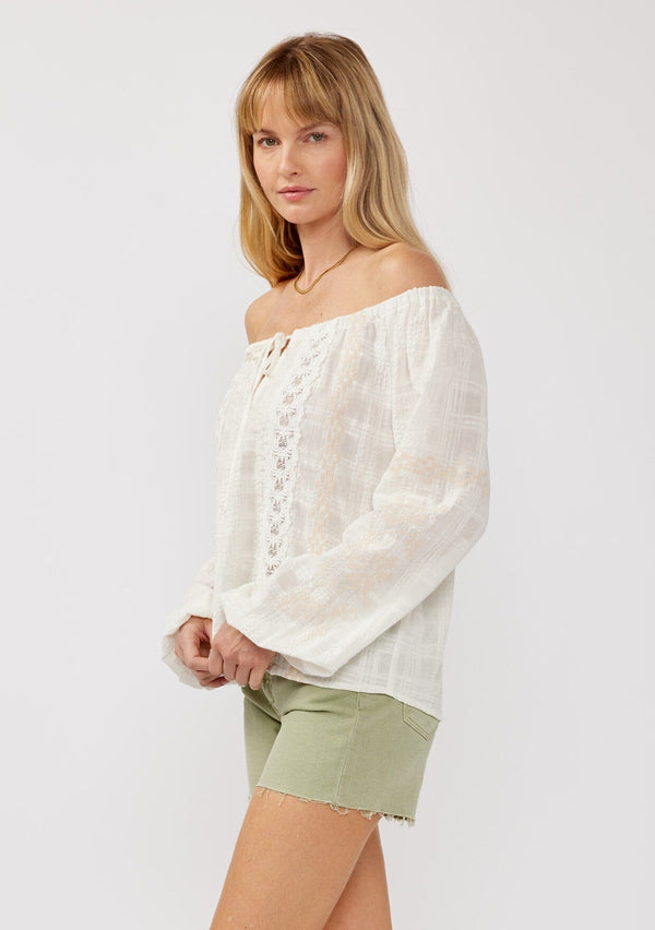 [Color: White/Taupe] A side facing image of a blonde model wearing an off shoulder top crafted in a textured plaid seersucker with an elastic neckline, tie neck detail, long sleeves, lace trim inserts, and contrast embroidery. 