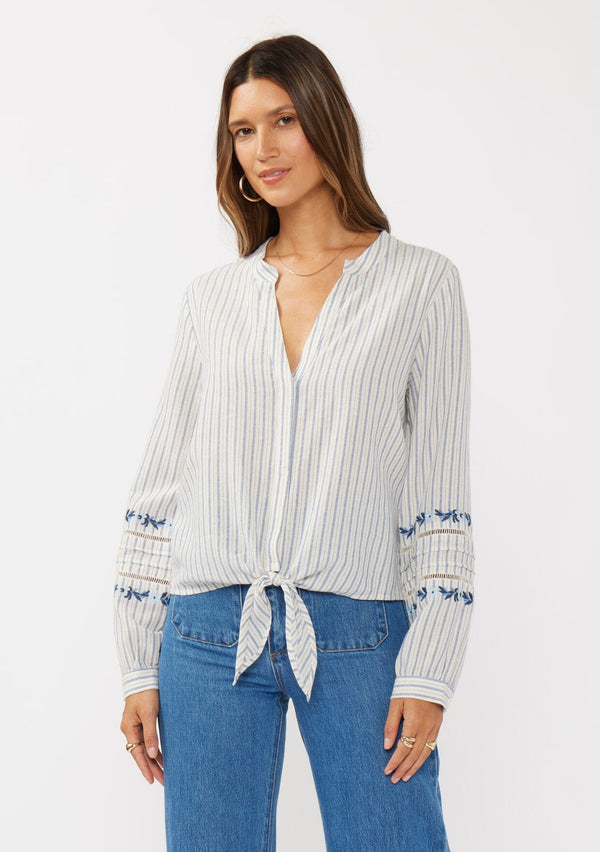 [Color: Navy/Natural] A front facing image of a brunette model wearing a blue yarn dye stripe blouse with a floral embroidery,  tie front, long sleeves with button cuffs, and a split v-neckline.